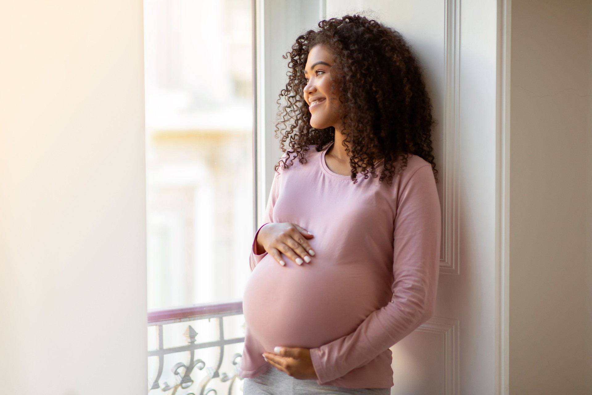 Black pregnant woman gently cradling belly while standing by window at home Black pregnant woman gently cradling belly while standing by window at home